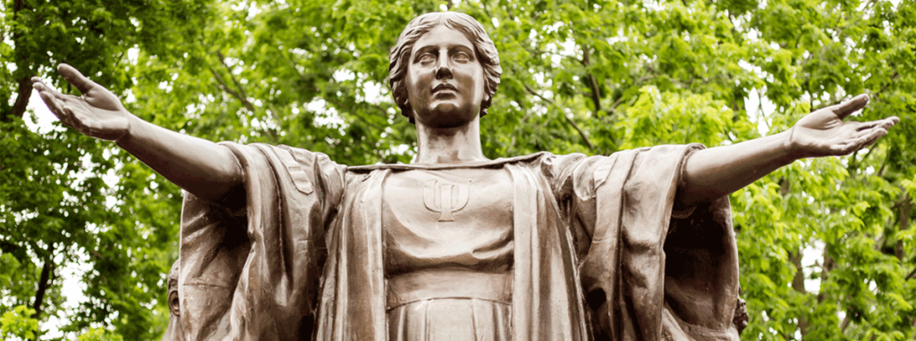 The Alma Mater Statue at the University of Illinois, with a backdrop of green summer leaves. Her arms are outstreched in welcome.