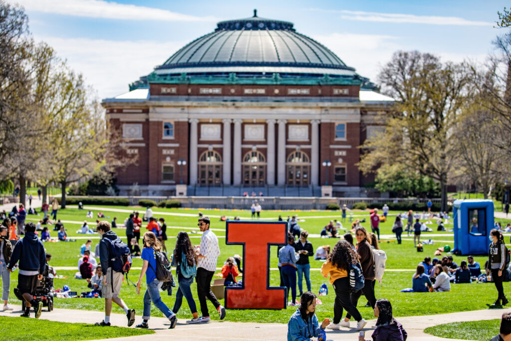 A spring break day on campus,. Students gather on the the grass in front of Foellinger Auditorium. A large Block I is in front of the building.